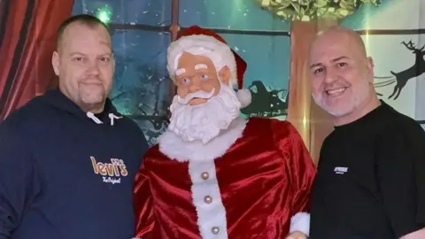 Rob Hardwick Two men standing either side of a model of Santa. The man on the left has short hair and a blue tshirt and the man on the right has a bald head and a black tshirt