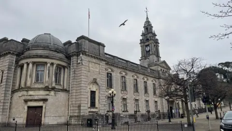 Torquay Town Hall, the headquarters of Torbay Council, seen on a cloudy day. It is an ornate building with a clock tower. There are black metal railings around it.