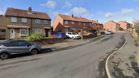 A quiet residential street lined with semi‑detached and terraced houses. The homes have brick exteriors, with many featuring red or brown roof tiles. Some houses have small front gardens or short driveways with low brick walls separating each property from the pavement. A few cars are parked along both the roadside and in driveways, including a grey car in the foreground and several others further up the street. The road itself curves gently uphill to the right, with a slightly worn but clean surface. The weather appears bright and clear, with blue skies and scattered white clouds. Trees and shrubs are visible along the right-hand side and near some of the gardens.