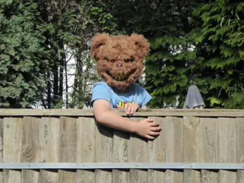 Verna Evans A child wearing a bear masks looks over a garden fence