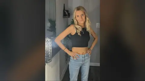 Carol O'Reilly Carol O'Reilly has long, blonde hair and is wearing a black crop top and faded blue jeans. She is standing by a mirror and is smiling at the camera 