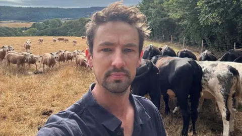 Chris Clements A selfie taken by a young man with brown hair standing in a field with cows and sheep. The man is wearing a dark blue polo shirt and looks worried.