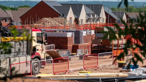 A housing construction site with a number of partially built houses. There is a lorry in the foreground carrying construction materials, and fencing surrounds the site. 