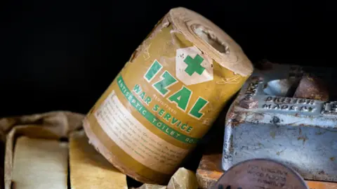 An old-looking roll of Izal toilet paper, the type that would have been used by the military in WW1. The paper cover around the roll says "Izal, Registered, War Service, Antiseptic Toilet Roll".