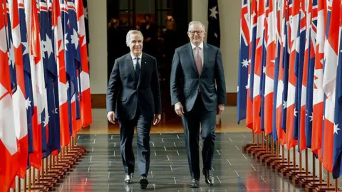 Getty Images Carney and Albanese are seen walking down a corridor that is lined with the flags of Australia and Canada. They are both wearing suits and are smiling. 