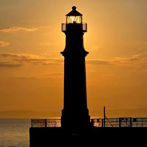 SCOTT MACKELLAR A lighthouse which is dark against the setting sun. The top of the lighthouse is glowing orange with the sun behind it. The sky is also orange with patches of dark grey and grey water below it.