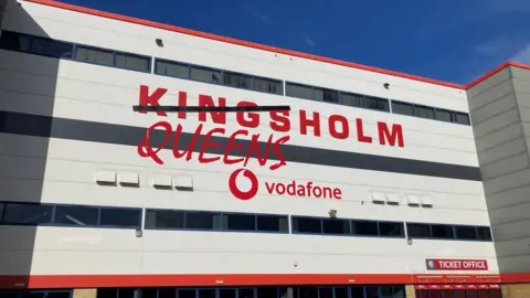 The four storey high Kingsholm stadium, home of Gloucester Rugby and Gloucester-Hartpury Women, has large red capital letters saying Kingsholm on the grey cladding. But a black line has been put across the first half of the word, covering Kings, and below it in a different font, is the word Queens.