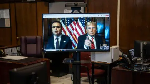 Donald Trump and his lawyer Todd Blanche (left) appear virtually on a computer desktop screen from Florida for his sentencing in his Manhattan hush money case. Trump has his finger pointing at the camera, with US flags positioned behind him and Blanche.