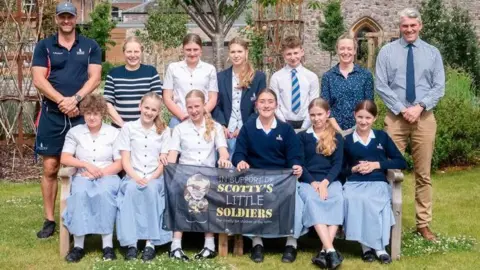Taunton Prep School Pupils in school uniform are sitting outside next to each other holding up a flag. They are smiling at the camera.