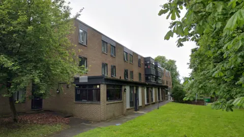 A three‑storey brick residential building with rows of windows and small balconies, set beside a paved walkway and surrounded by grass and leafy trees on a quiet, green campus‑like setting.