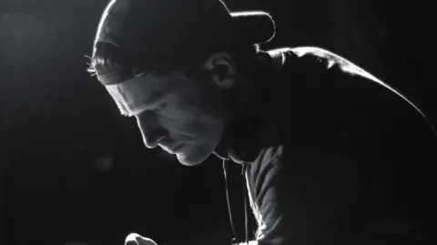 Kelly-Anne Bond A side profile of Arthur's face in black and white, silhouetted by light. He is leaning over a camera wearing a baseball cap backwards