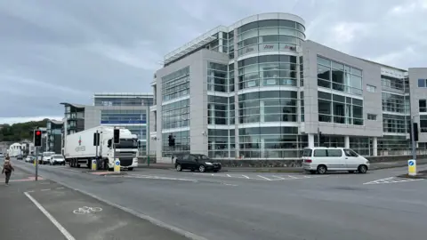 BBC A grey building with blue windows with a traffic light controlled junction in front. An articulated lorry is waiting at the lights and two cars are turning left into Admiral Park. 