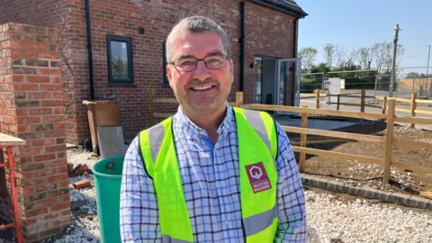 Chris Carr stood on a construction site where a house is being built. He has short grey hair and facial stubble. He is wearing a blue, grey and white checked, long-sleeve shirt and a yellow hi-visibility vest. He is also wearing rectangle, black-rimmed glasses. He is smiling at the camera.