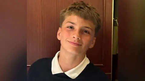 Josh Travis is pictured smiling at the camera. He has medium length brown hair which is short at the sides. He is wearing a white school polo shirt and dark navy jumper
