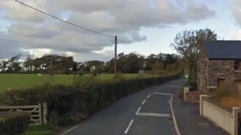 Google The road leading away from the junction with St Judes. There is a green field and hedge on the left and a wall and stone built building on the right.