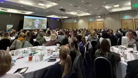 Lots of people sitting around tables looking at the speaker on a screen for the conference.