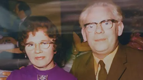 Betty and Cyril looking at the camera. They both wear glasses. Betty is wearing a purple top and Cyril is wearing a suit.