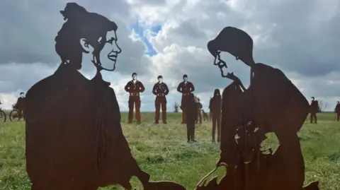 Steel statues of silhouettes on Salisbury Plain. Three soldier statues can be seen in the distance. Statues of two women sat on the grass talking can be seen in the foreground of the image