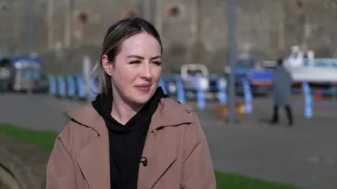 Bekki has blond hair, tied back in a middle parting. She has a wooly neutral coloured scarf on and a brown trench coat. Behind her is a harbour.