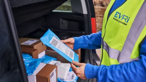Evri courier in blue sweatshirt and high-vis tabard is seen from waist to neck. He holds a small parcel wrapped in blue plastic with a white label. He stands at the open boot of a grey van filled with parcels.