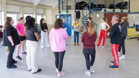Johan Persson A woman wearing a yellow scarf speaks to a group of women and one men standing in a circle. In the background is a metal spiral staircase.