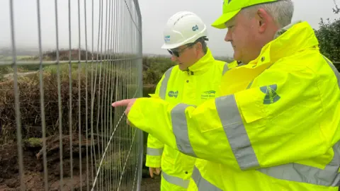 Two men in SWW branded high viz jackets and hard hats are standing behind a metal fence looking at a muddy area which the man in the foreground is pointing to.