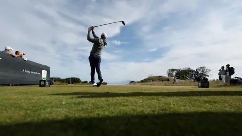 Getty Images Scottie Scheffler pictured holding his driver aloft after teeing off. A small number of fans on either side follow the flight of the ball. Scheffler is wearing a grey hoodie, navy trousers, a white hat and white shoes.