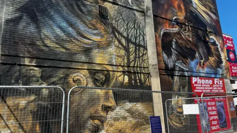 A mural on the side of a concrete wall, on the left a design of the right side of a statue's face coloured in yellow and grey, on the right an image of an eagle with wings spread holding an oil lamp. There is a phone repair advertisement in the bottom right corner and scaffolding in front of the mural.