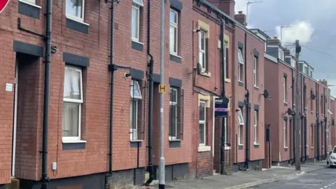 BBC Terraced redbrick houses, Some with black grills over the windows. One has a purple 'Let By' sign at the front.