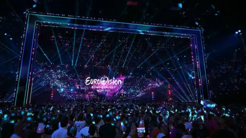 Getty Images A huge stage that is dark, with a screen that says "Eurovision song contest, Basel 2025". There are fluorescent lights of blue, purple and pink. A crowd of thousands stands in front of the stage, reflected with blue light. Many have their phones out.