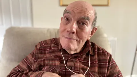 Alex Pope/BBC Clive, sat on a sofa, wearing a brown checked shirt, with a cord round his neck. He has a bald head and grey hair at the sides. He is looking towards the camera. 