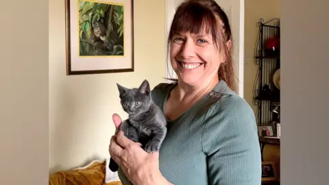 Lisa Burtenshaw Lisa Burtenshaw is pictured holding a small grey kitten. Lisa is wearing a teal-coloured v-neck top. She has brown hair that is tied in a ponytail and she is smiling widely and looking directly at the camera. She is in a front room.