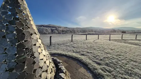 The Cairn Distillery Frost covers the ground in the morning near the Cairngorm mountains. A sculpture also glistens, covered in ice. In the distance, the mountains Carn na loinne and carn na coinnse xan be seen.