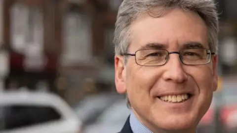 Mr Foster is smiling at the camera in a close-up photo. He has glasses on and a blue shirt and dark suit jacket. He is standing outside on a street.