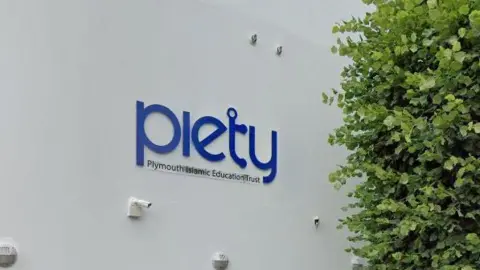 Outside the Plymouth Islamic Education Trust's building in Plymouth. A blue sign for the charity is on a white wall. A CCTV camera is on the wall. A large tree is on the pavement outside the building along with a parked silver Ford Focus.