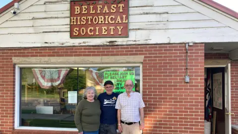 Sean Drysdale Sean Drysdale stands between a man and woman who look to be in their 50s or early 60s in front of a brick building with the sign Belfast Historical Society on it 