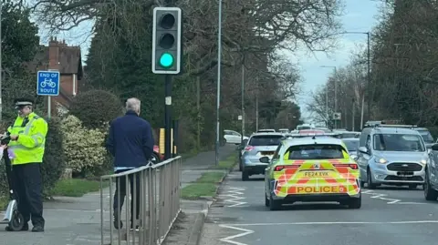 Ali Bulley A picture of a busy single-lane road taken by a driver, with a police car in front. A police officer in high-vis yellow jacket is chatting to someone on the side of the road. A pedestrian crossing with traffic lights can be seen.