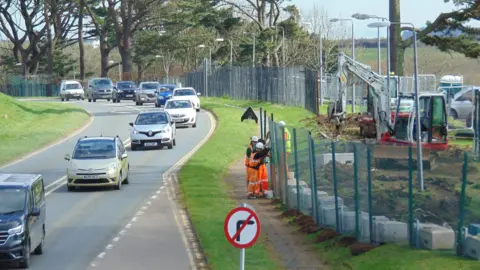 Contractors wearing high-viz clothing work on installing the new fencing next to a busy road. There is a mini-digger in action in a gap between sections of green fencing. There are also trees lining a section of the fence.