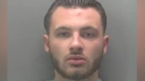 Cambridgeshire Police Police mugshot of Chas Corrigan. He is wearing a grey top and has brown hair swept back and a beard.