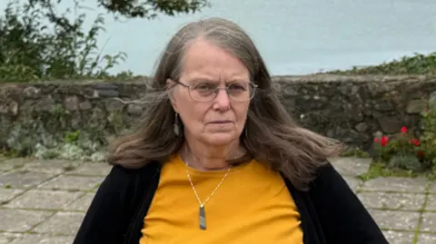 Morfudd Mill is standing on a paved area with trees and the sea in the background. She is wearing a mustard yellow top, black cardigan and green pendant. She has long grey and brown hair.