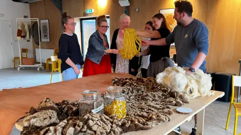 A group of people stand around a table. They are looking at yellow wool, there are other items on the table.