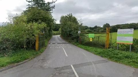 Google Maps The road leading into Sherborne Recycling Centre. There are trees and bushes either side of the road. There is a sign to the right of the image. There are yellow gates, which are open, in the foreground.