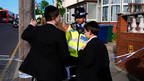 PA Media Two children speak to a police officer in front of a police cordon at the scene