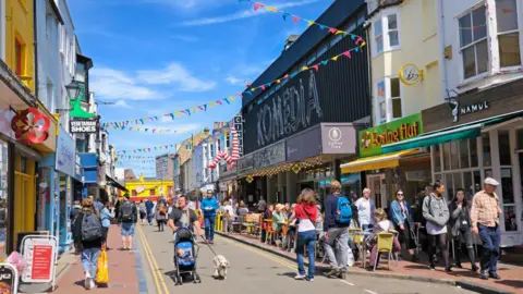 Getty Images A vibrant city centre area with bunting between the buildings and people on the streets. The sky is blue.
