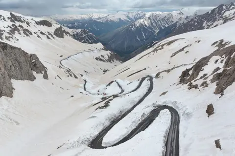 Anadolu via Getty Images Aerial view of a winding mountain road cutting through deep snow on the Van-Bahçesaray highway in Van, eastern Turkey, where snowplow crews and vehicles work along sharp bends amid steep, snow-covered slopes and rugged peaks under a cloudy sky.