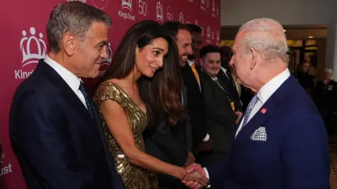 Amal Clooney, dressed in a golden dress, shakes hands with a navy blue-suit wearing King Charles as her Hollywood star husband George, also in a dark blue suit, beams proudly at her right hand side during a line up of famous names meeting the monarch