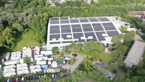 Bottle Yard Studios An aerial view of Bottle Yard studios showing a large warehouse roof covered in solar panels and surrounded by trees, there appears to be seven rows of trailers in the bottom left corner of the frame.
