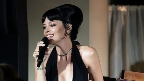 Getty Images Lily Allen is singing into a mic. She has a black low neck dress on, her black hair is in an up-do with a beehive. She has a silver necklace on. 