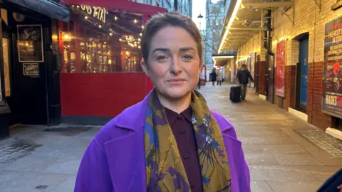 BBC Ros Morgan, wearing a purple jacket and floral scarf, sees at the camera. SHe has short hair and a neutral expression