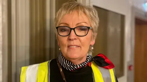 Eileen is looking directly at the camera. She has blond hair and is wearing black-framed glasses. She has a yellow high visibility jacket on top of a black jumper. 